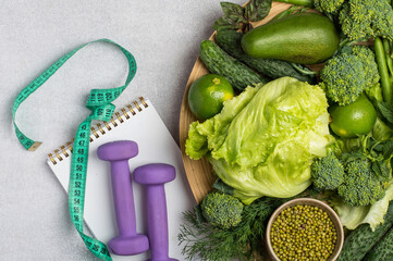 Fresh green organic vegetables in assortment, dumbbells, tape measure on light background. Healthy lifestyle, weight loss, fitness, detox, flat lay.