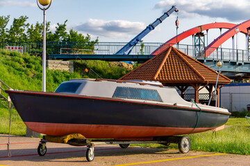 Motorboat placed on a transport trailer, parked in a port area next to a wooden shelter. In the background, an arch bridge and a construction crane are visible. Scene from a harbor environment.