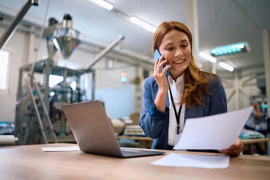 Happy female business owner going through paperwork while talking on cell phone at factory production plant.