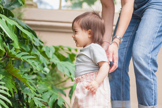 A baby girl observes green leaves up close, supported by her mother's hands as she takes her first steps. A tender moment of discovery and care in a natural setting.