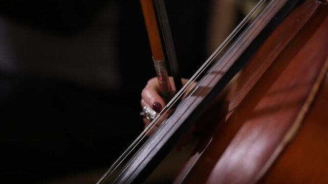An elegant close-up of a cellist's hand bowing strings. The shot shifts to the bridge, showing detailed, skilled technique during a classical music performance with beautiful bokeh.