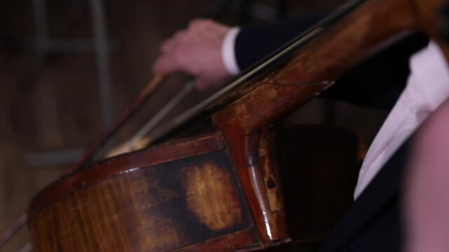 An elegant close-up of a cellist's hand bowing strings. The shot shifts to the bridge, showing detailed, skilled technique during a classical music performance with beautiful bokeh.