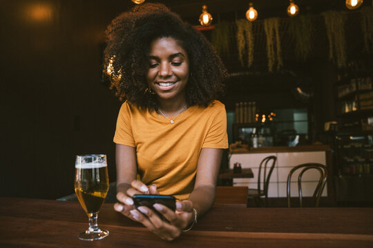 Smiling young woman using smartphone while sitting with beer in cozy cafe