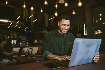 Fototapeta premium Smiling young man typing on laptop while working remotely in cafe