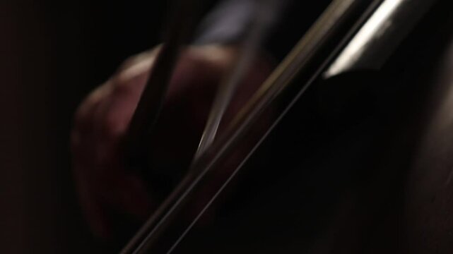 An elegant close-up of a cellist's hand bowing strings. The shot shifts to the bridge, showing detailed, skilled technique during a classical music performance with beautiful bokeh.