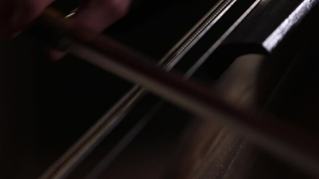 An elegant close-up of a cellist's hand bowing strings. The shot shifts to the bridge, showing detailed, skilled technique during a classical music performance with beautiful bokeh.
