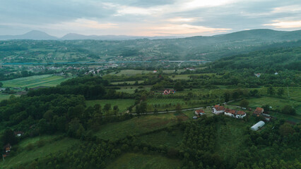 Fototapeta premium Aerial view of green rural landscape with red-roof houses surrounded by fields and orchards in Serbia at sunset