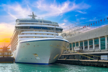A modern white cruise ship sits docked at a pier. The ship is tied with thick yellow ropes. A...