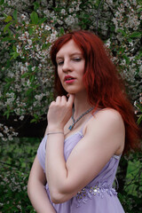 red-haired girl with curly hair walks in the park. Girl in a smart dress smiles. Woman poses against the background of blooming yellow dandelions and white flowers in a turquoise dress