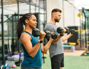 Portrait of a young black woman and white man coach or trainer exercising in a gym, lifting weights, dumbbell equipment, healthy lifestyle and strength exercise at fitness club concepts