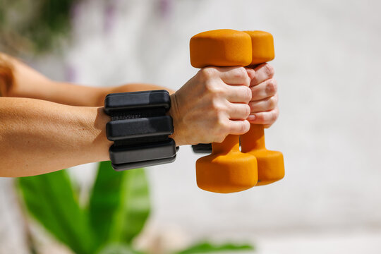 Close-up of female hands gripping orange dumbbells and wearing black wrist weights, symbolizing strength, fitness, and motivation during a focused outdoor workout on a sunny day.