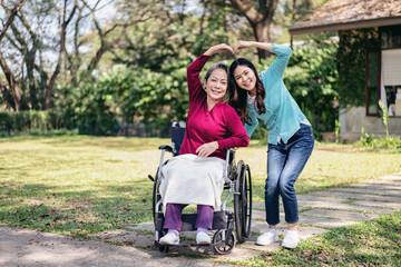 Asian Adult Woman and Older Asian Woman in Wheelchair Making Heart Shape with Hands in Garden. Caregiver and Elderly Care Concept. Family Love in Nature.