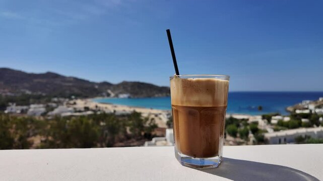 View of a delicious greek iced coffee, also known as cafe frapp&eacute;e and the beautiful Mylopotas beach in Ios Greece in the background