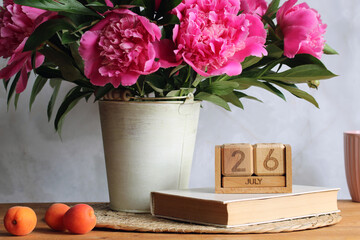 Pink peonies in metal vase, wooden calendar, book and apricots on table celebrating july 26th