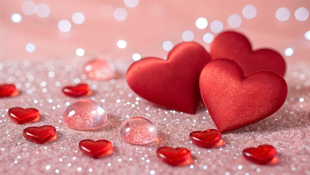Red hearts and decorative gems are arranged on a pink glittery surface for valentines day