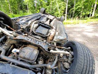 Vehicle is flipped onto its roof on forest gravel road in wooded area, undercarriage damage.