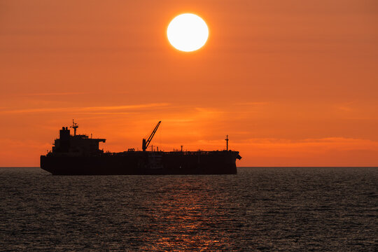 Dark silhouette of a large sea tanker with a crane on deck in the calm waters of the Baltic Sea against a majestic orange sunset. - Powered by Adobe
