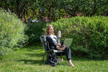 Relaxed businesswoman confidently working on laptop while hydrating on grass in park. Confident...