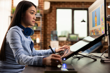 Asian graphic designer holding stylus pen, using digital art pad to modify images on computer screen. Skilled woman focused on retouching photos and refining design details for content production.