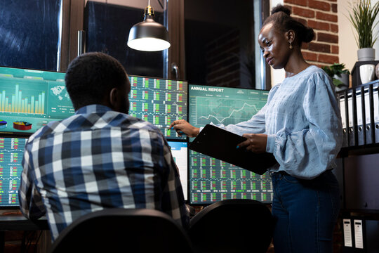 Two financial analysts collaborate at desk in office, reviewing stock market information on multiple monitors at night. Female manager with a clipboard, stands and explains forex trading to employee.