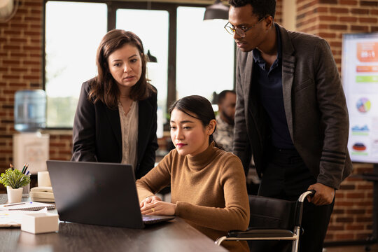 Diverse team of colleagues collaborating in modern workspace, sharing strategy ideas in brick wall office. Woman in wheelchair discussing with her coworkers and checking business analytics on laptop.