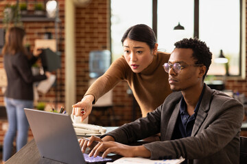 Female analyst points at laptop screen to explain market analysis to a african american coworker....
