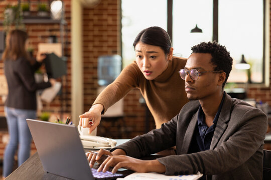 Female analyst points at laptop screen to explain market analysis to a african american coworker. Black man listens attentively as asian manager addresses financial errors in the presentation.