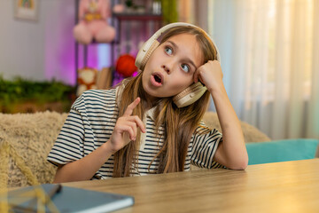 Young child girl sitting at table on home sofa listens to an audio fairy tale in wireless headphones. School kid excitedly opens eyes smiles with wonder imagining magical events and curious moments