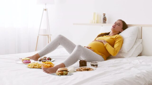 A woman in her third trimester rests comfortably on a bed, gently holding her belly. She is surrounded by an assortment of comfort foods, feeling bad after eating junk unhealthy food