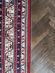 Close-up of a traditional patterned rug with red, navy, and cream tones next to a dark herringbone wood floor, showcasing rich texture and interior contrast.