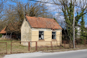 Small abandoned rural house with a red tile roof and broken windows sits behind a rusted fence, surrounded by overgrown grass and leafless trees under a blue sky, showing signs of decay