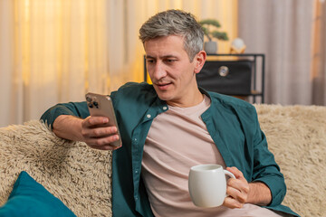 Young man at home starts morning sitting on sofa with tea, reading smartphone with calm focus. Caucasian guy looks peaceful, focused on news and relaxed, enjoying warm drink in quiet home moment.