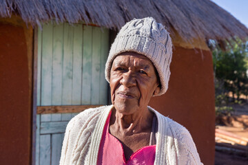 village african old woman standing in the yard in front of a hut with thatched roof, wearing a beanie