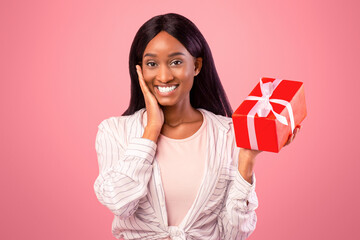Portrait of suprised African American woman with wrapped gift box posing on pink studio background....