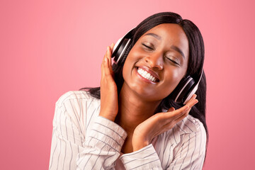 Fototapeta premium Portrait of pretty African American woman wearing headphones and listening to beautiful music on pink studio background. Lovely black lady enjoying her favorite melody with closed eyes