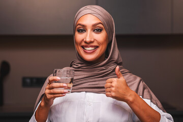 Muslim Woman Holding Glass Of Water Gesturing Thumbs Up Approving It Posing Smiling To Camera In Modern Kitchen Indoor, Wearing Hijab. Arab Lady Advertising Mineral Water. Healthy Hydration Concept