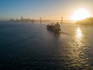 Obraz premium Cargo ship sails into San Francisco Bay at sunrise, passing the Bay Bridge and city skyline, facilitating global trade in USA.