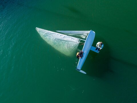 Aerial view of two sailors in life jackets righting their capsized dinghy during a sailing lesson or practice on the water. Point Richmond, Richmond, Caifornia, United States