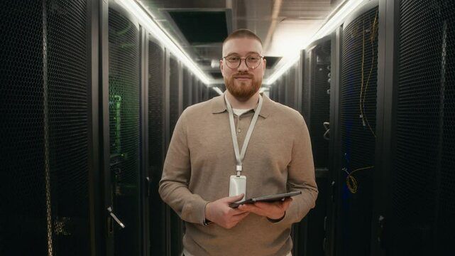 Wide portrait of cheerful male Caucasian network engineer wearing badge looking at camera in data center