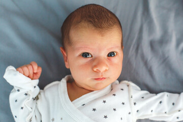 Newborn baby lying on a soft gray bed in white star-printed clothes, looking directly at the camera...