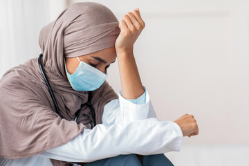 Sad arab woman doctor having difficult day at clinic, wearing protective face mask, sitting next to...