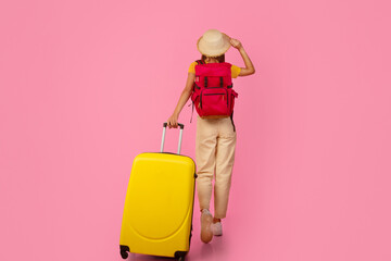 Fototapeta na wymiar Back view of unrecognizable woman backpacker walking with yellow suitcase on pink studio background, touching her summer hat, full length photo, copy space. Female tourist with backpack and luggage