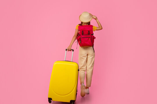 Fototapeta Back view of unrecognizable woman backpacker walking with yellow suitcase on pink studio background, touching her summer hat, full length photo, copy space. Female tourist with backpack and luggage
