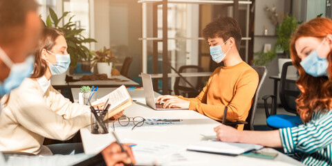 Fototapeta premium Prevention And Precautions. Group of diverse international students or employees wearing protective surgical masks using laptop computers, studying and working, people reading and writing in office