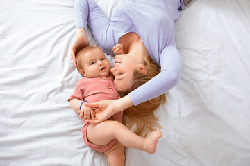 Adorable baby boy and loving blonde mother laying on bed head to head, top view. Joyful young woman playing with her cute little kid on bed, laying and bonding together at home