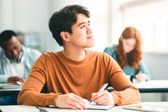 Portrait of smiling chinese male university student holding pen writing in notebook doing examination or quiz test from teacher sitting at desk, attending classroom lesson, looking aside and thinking