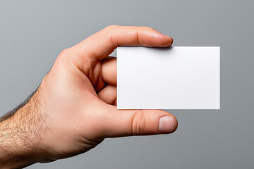 Obraz premium Close-up of a male hand holding a blank business card against a neutral gray background, perfect for showcasing branding, contact details, or professional identity mockups