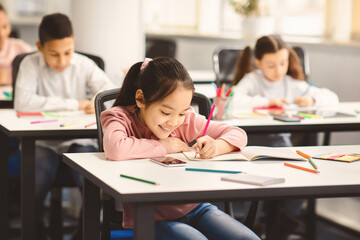 Portrait of small smiling asian girl sitting at table in classroom at primary school or kindergarten, writing or drawing in notebook. Reopening and return back to school after coronavirus quarantine