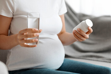 Prenatal Vitamins. Unrecognizable pregnant woman holding blank plastic jar with pills and glass of water, using supplements for healthy pregnancy, sitting on couch at home, cropped image, closeup