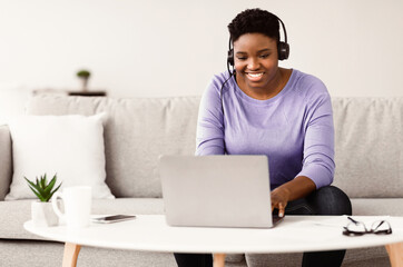 Telecommuting Concept. Smiling curvy african american woman sitting on the sofa and working online on laptop, wearing headset. Young black female using computer at home office, typing on keyboard